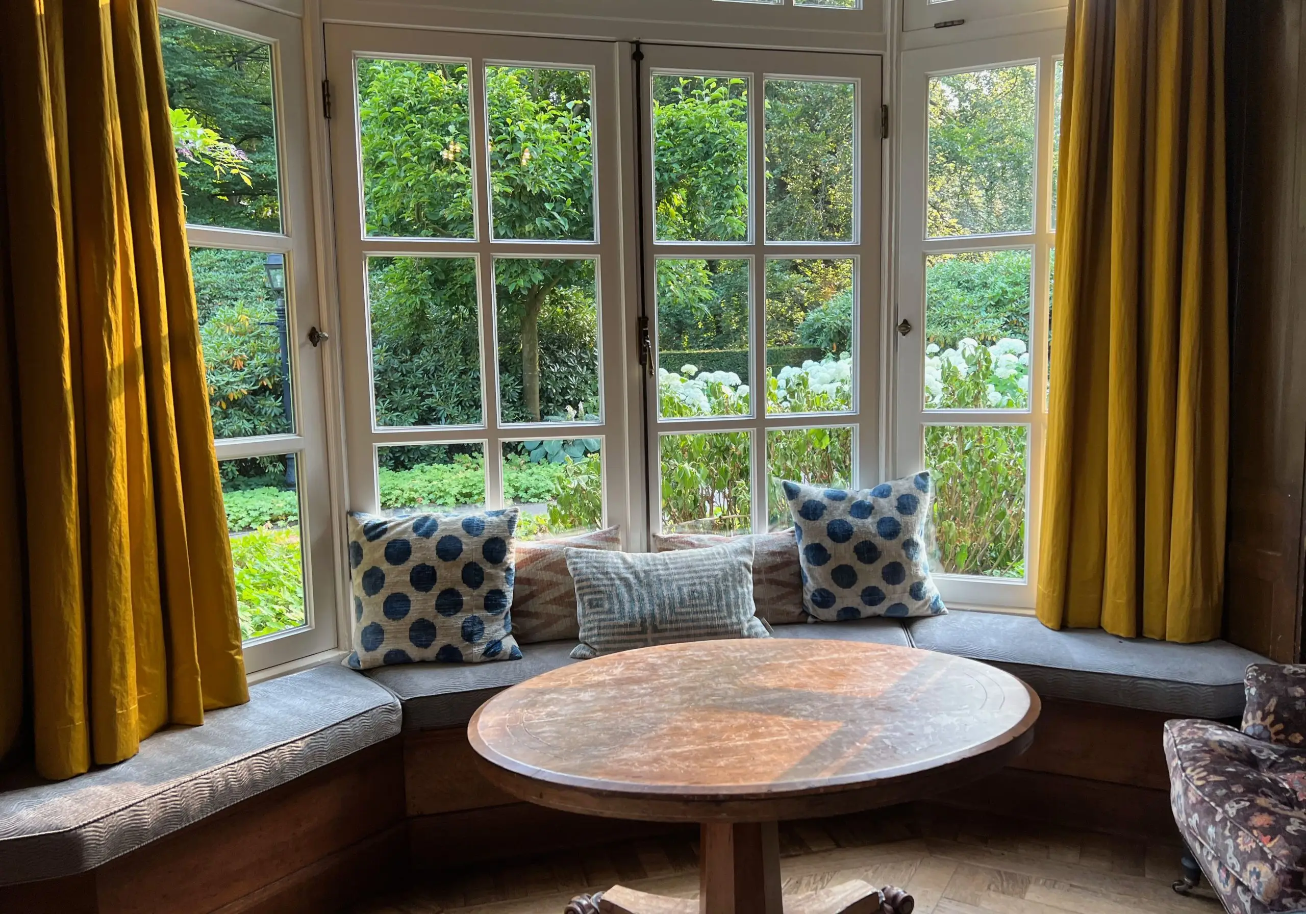 Wooden table near cozy bay window seat with cushions in room. Interior design